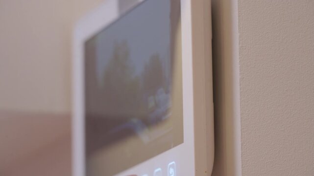 A modern white intercom system is mounted on a textured wall, its screen displaying a blurred outdoor scene, hinting at communication and connection with the external environment.