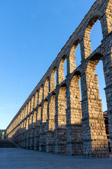 Fototapeta premium The massive Roman Aqueduct is bathed in golden sunset light in Segovia, casting long dramatic shadows across the plaza. The ancient stone arches loom over the urban landscape.
