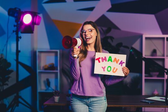 Young female blogger holding a thank you sign and megaphone in a vibrant indoor setup with neon lighting