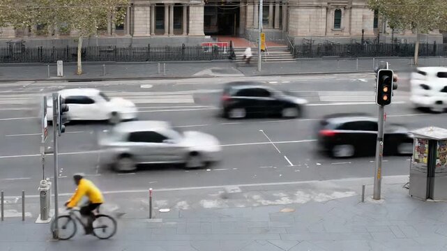 A cyclist in a bright yellow jacket navigates a busy city street as cars rush by, highlighting the contrast between slower pedal power and fast urban traffic in motion.