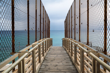 A symmetrical view down a weathered wooden pier that extends over the turquoise sea towards an...