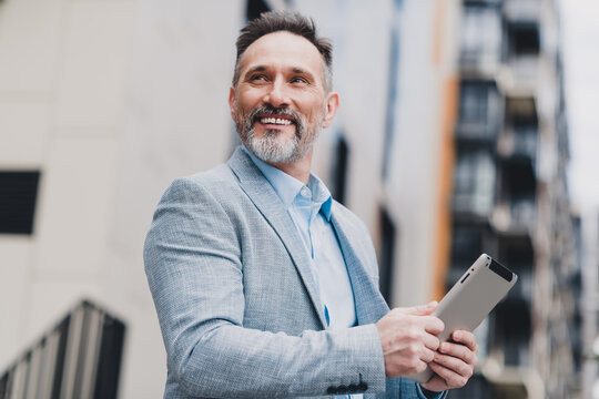 Confident mature businessman stands on a city street holding a tablet smiling in a modern outfit conveying style and success in urban professional life