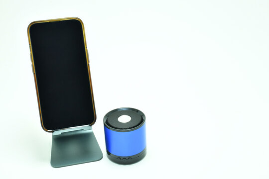 Tel&eacute;fono m&oacute;vil sobre una base de metal, al lado una bocina inal&aacute;mbrica sobre un fondo blanco, espacio para texto al lado derecho y pantalla. Toma horizontal.