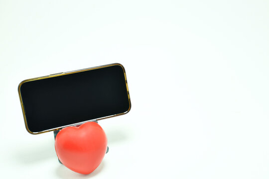 Tel&eacute;fono m&oacute;vil sobre una base, abajo un coraz&oacute;n rojo con fondo blanco. Toma horizontal, espacio para texto al lado derecho y pantalla. Uso de la tecnolog&iacute;a para el cuidado de la salud.