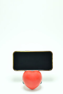 Tel&eacute;fono m&oacute;vil sobre una base, abajo un coraz&oacute;n rojo con fondo blanco. Toma vertical, espacio para texto en la parte superior y pantalla.