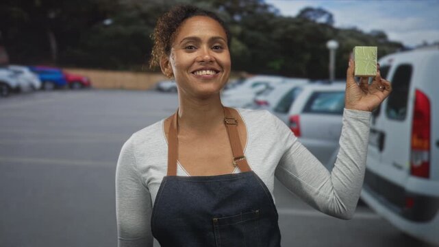 Woman holding a small green box and smiling in a street parking area with cars visible; small business pride.