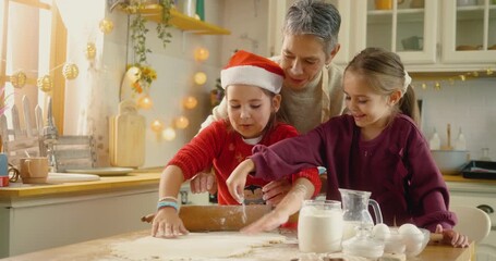 Grandmother teaching granddaughters how to roll dough while baking Christmas cookies together. Joyful family bonding in cozy kitchen filled with laughter and holiday spirit.