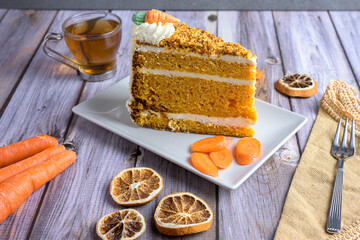 Slice of homemade carrot cake with cream cheese frosting and tea on rustic wooden table, decorated with dried orange and carrots, cozy autumn dessert concept