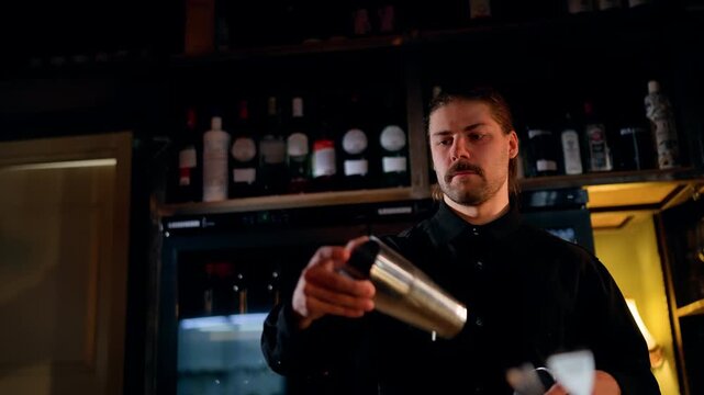 Focused bartender performing cocktail flair behind a dimly lit bar, tossing a metal shaker and preparing drinks amid shelves of bottles, warm ambient lighting, and glassware.