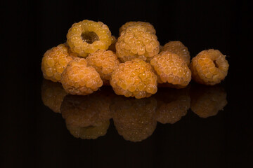 Close-up of a group of ripe yellow raspberries against a glossy black background. Bright overhead lighting highlights the berries' texture and creates a symmetrical reflection.