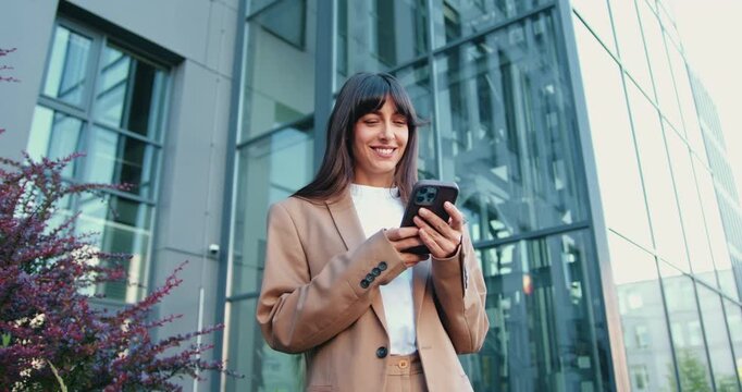 Young Caucasian businesswoman standing outside and smiling while using smartphone, with modern glass office building in the background