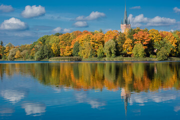 A landscape where the pointed spire and the top of an ancient tower rise above a dense line of autumn trees. The vibrant foliage is tinged with rich yellow and orange tones.