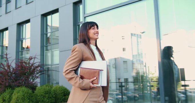 Side view of a young Caucasian woman confidently walking near a modern glass office building, smiling as she carries a laptop and a notebook in her hands. Joyful and determined on her way to work