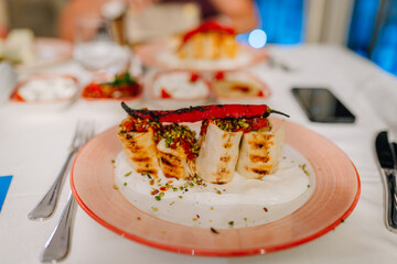 Close-up of a plated dish featuring grilled wraps with pistachio topping, creamy sauce, and a red pepper garnish on a restaurant table.