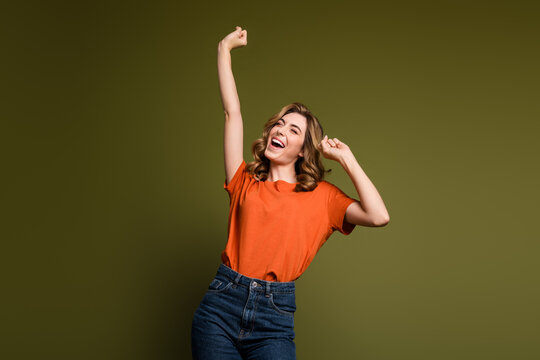 Joyful young woman with curly blonde hair in orange t-shirt posing cheerfully against a stylish khaki background