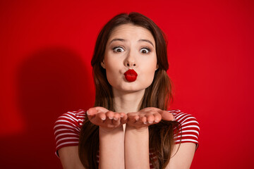 Charming young woman blows a kiss while wearing red lipstick against a vibrant red background,...