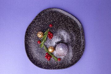 Top view of a Christmas dessert and golden bells on a black plate on a violet background.