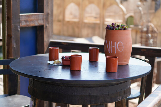 Terracotta jug labeled "VINHO" filled with grapes, surrounded by cups on a round table in a rustic setting. Wine culture, Portugal.