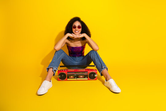 Energetic young girl with curly hair wearing a purple sequin top and jeans sits against yellow backdrop posing with a red boombox - Powered by Adobe