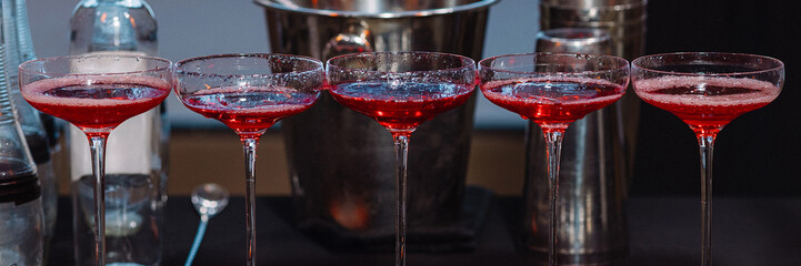 Row of five red cocktails in coupe glasses on black table with shaker, bottle and ice bucket. Indoor bar setting. Concepts: cocktail party, celebration, nightlife, alcohol menu, mixology,bar service.