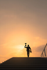 Fototapeta premium Triumphant ascension: Silhouette of a businessman reaching the summit, holding a trophy against a vibrant sunset. Success achieved!