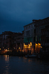 A captivating nighttime view along a Venetian canal. Warm light spills from the windows of historic buildings reflecting on the water's surface, creating an enchanting atmosphere. 
