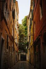 Narrow alleyway recedes into historic Venice, flanked by aged buildings with weathered shutters. Sunlight illuminates textures and creates depth within the charming scene, evoking a sense of mystery.