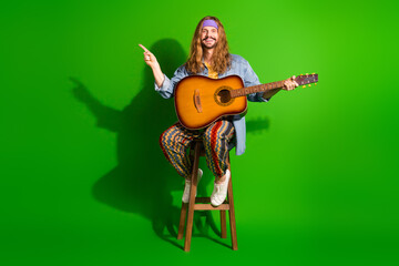 Fototapeta premium Hippie-style musician on wooden stool with guitar, green background, finger pointing, vibrant fashion and retro vibe