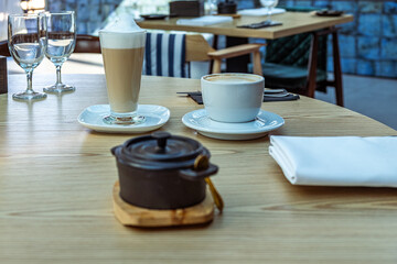 Coffee and dessert served on a wooden table in a modern cafe, featuring a latte, cappuccino, and a small pot of chocolate, creating a cozy dining atmosphere