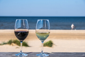 Two elegant wine glasses filled with red and white wine sit on a wooden table overlooking a serene beach, capturing a moment of relaxation and enjoyment by the sea