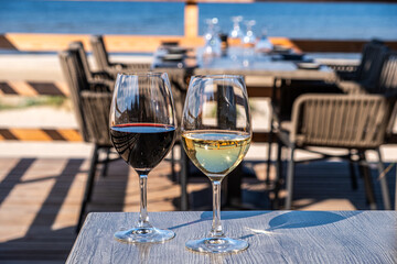 Two elegant wine glasses filled with red and white wine are placed on a wooden table, showcasing a beautiful outdoor dining setting with a serene waterfront view