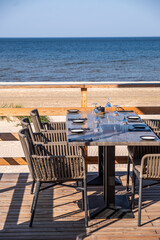 Outdoor dining table set for a meal, surrounded by stylish chairs, overlooking a serene beach and calm ocean, creating a perfect atmosphere for relaxation and enjoyment
