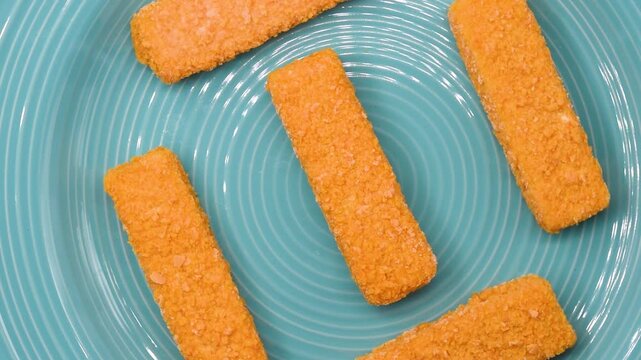 Frozen breaded cod fish fingers rotating on a blue plate. Ready-to-cook seafood product ideal for fast meals, kids&rsquo; food, or frozen food advertising.