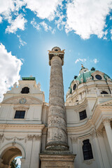 Vienna Karlskirche column soars against bright blue sky, sunlit baroque reliefs in dramatic low angle, historic grandeur, ideal for travel ads and editorials.