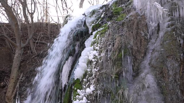 A frozen waterfall on a mountain river. The Bigar waterfall below Stara Planina is bound by ice. details of ice stalactites and other ice formations. Unfrozen falling water flows between the ice
