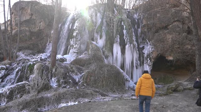 A frozen waterfall on a mountain river. The Bigar waterfall below Stara Planina is bound by ice. Unfrozen falling water flows between the ice. Tourists approach the waterfall and look at it.