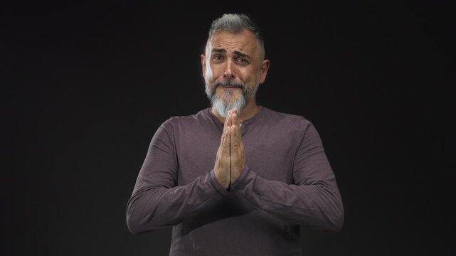 Middle aged man with hands pressed together in a black studio, palms touching and pleading expression, bare hands visible; pleading apology.