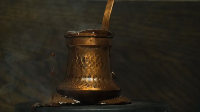 A coffee drink boils over from a copper cezve with foam, spilling onto a hot stove burner during brewing. The overflowing coffee hisses, bubbles, steams, and splashes drops of hot black liquid.