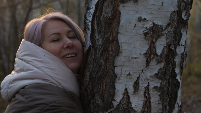 A happy Caucasian woman, 45 years old, hugs a tree in a forest or park, the concept of the unity of man with nature, a woman walking in the forest in autumn