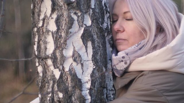 A beautiful middle-aged Caucasian blonde woman stands and hugs a tree while walking in an autumn forest at MLM Park, a concept of state of mind and communication with nature.
