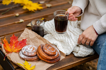 Cozy autumn picnic with a woman in a knitted sweater holding a glass cup of coffee next to cinnamon rolls and colorful maple leaves on a wooden bench