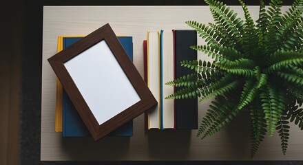 A top view captures books, a picture frame, and a lush green fern arranged on a light wood surface. The setup suggests a calm, aesthetic workspace or home decor.