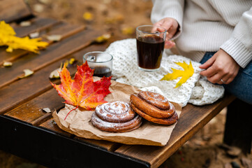 Partial view of woman holding cup of coffee on wooden bench with cinnamon rolls and fall leave