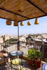 A cozy terrace in Morocco welcomes relaxation with a small table and chairs surrounded by lush plants. The view captures rooftops under a bright blue sky, inviting visitors to unwind