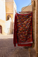 Brightly patterned textile drapes across a narrow street in El'Jadida, creating a vibrant focal point against the sunlit stone buildings. The scene captures local culture on June 13, 2021