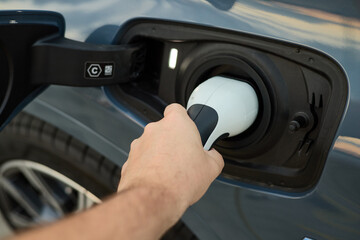 A person connects a charging cable to an electric vehicle at a public charging station. The location features a sleek design and well-maintained surroundings in broad daylight.
