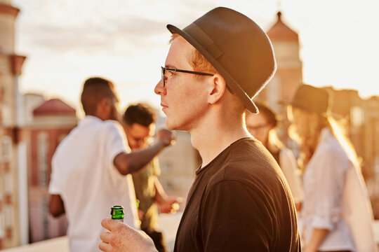 Profile portrait of young stylish Caucasian man in bowler hat and glasses standing on rooftop with beer and looking into the distance, friends hanging out in the background - Powered by Adobe