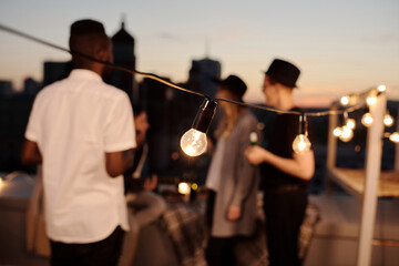 Fototapeta premium Close-up view of decorative lightbulbs illuminating rooftop where defocused young people standing and chatting on summer evening