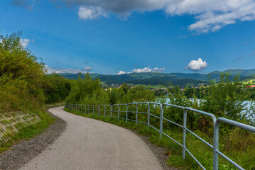 Fototapeta premium Winding path follows the lake’s edge with protective railings and panoramic mountain view ahead. Perfect scene for cycling, walking, or promoting outdoor lifestyle and eco-friendly travel.