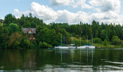 Peaceful sailboats rest at a small marina surrounded by lush green forest. The tranquil water reflects trees and white clouds, creating a serene summer atmosphere perfect for relaxation and nature tra